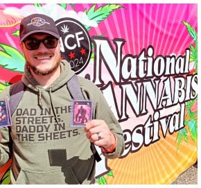 Image of Stephen Chappell in a black baseball cap with the Legendary Leafies logo holding up a trading card and standing in front of a colorful banner for the National Cannabis Festival.