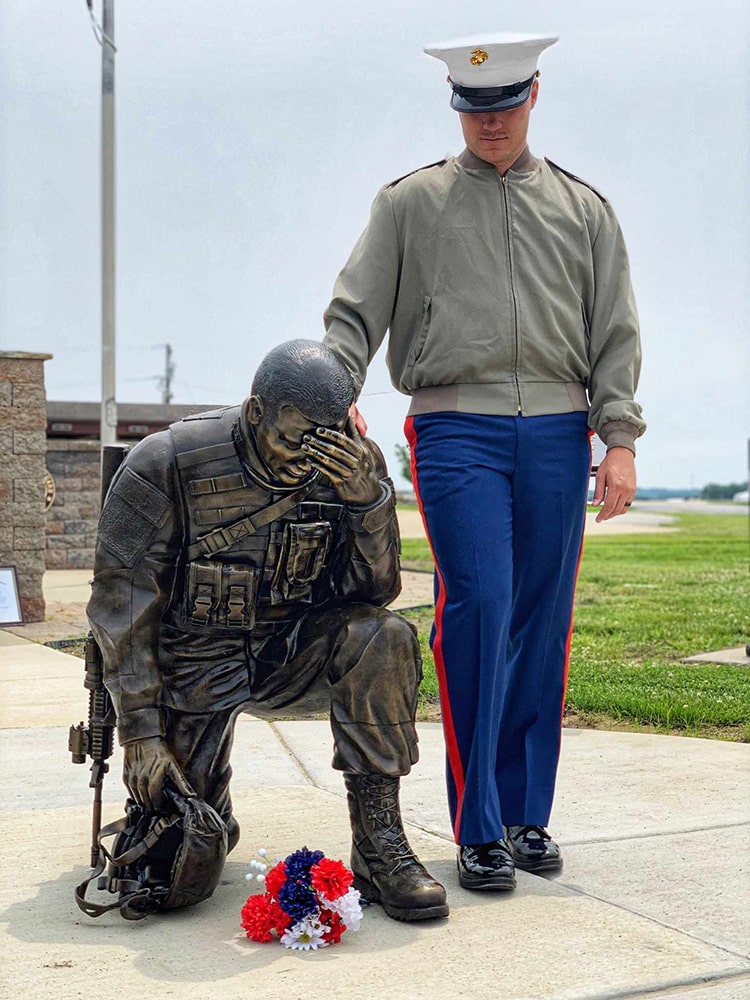 Stephen Chappell dressed in his Marine Corps uniform standing with a bronze statue of a grieving soldier with red flowers in the foreground.